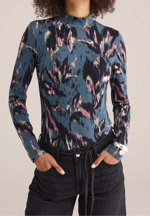 Kobieta ubrana w długi rękaw z abstrakcyjnym wzorem w kolorach niebieskim, różowym i czarnym, włożonym do czarnych, wysokich spodni z rękami w kieszeniach.