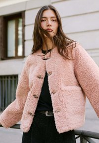 Helle rosa, flauschige, strukturierte Jacke mit rundem Kragen, Fronttaschen und Knebelverschlüssen; getragen über einem schwarzen Oberteil und dunklen Hosen.