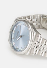 Montre en acier inoxydable argenté avec un cadran bleu clair, des index horaires minimalistes et une finition brossée. Elle est dotée d'un bracelet à maillons et d'une couronne.