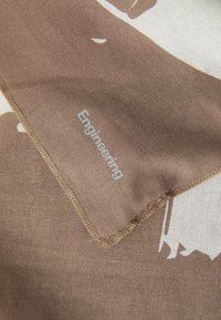 Brun stoffskjerf med en silketrykt design i hvitt, avsluttet med beige sydd kanter og ordet "Ingeniørfag" i grått.