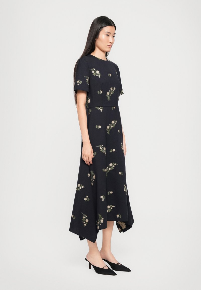 Robe florale bleu foncé à manches courtes, avec un ourlet asymétrique et de petits motifs floraux en vert clair et crème. Associée à des talons noirs.