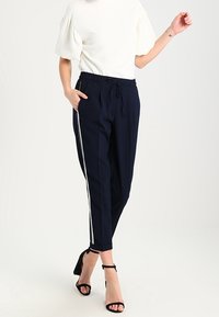 Granatowe spodnie dresowe z białymi paskami po bokach, elastycznym pasem i kieszeniami. W zestawie z białą bluzką i czarnymi sandałami na obcasie.