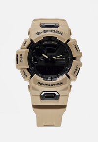 Montre G-SHOCK beige avec un cadran noir, dotée d'un boîtier en résine, d'un affichage numérique, de gros boutons, et marquée "PROTECTION" et "LIGHT".