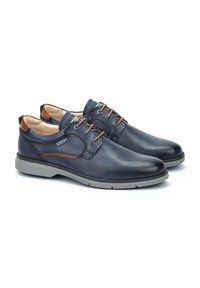 Chaussures en cuir bleu foncé avec des accents marron. Design à lacets et semelle en caoutchouc gris clair, présentant une texture lisse et des détails minimalistes.