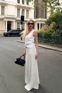 Weißer strukturierter Stricktop mit V-Ausschnitt und passendem langen Rock. Die Modelführt eine kleine schwarze Handtasche. Städtische Straßenkulisse.