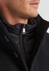 Schwarzer Außenmantel mit einer strukturierten Wolloberfläche, ausgestattet mit einem hohen Kragen und einer darunterliegenden schwarzen Pufferjacke mit Reißverschluss. Sichtbares silbernes Reißverschlussdetail.