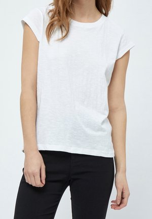 Femme portant un t-shirt blanc à manches courtes uni et un pantalon noir, debout devant un fond clair uni.