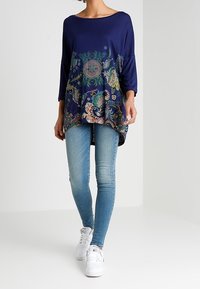 Tunique bleu marine avec un imprimé floral et géométrique coloré au bas, associée à un jean slim bleu clair et des baskets blanches.