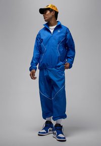 Blå träningsdräkt med vita inslag, jacka med dragkedja, elastiska ärmslut och byxor med lös passform. Kombineras med blå och svarta sneakers samt en gul keps.