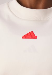 Bílá mikina s texturovaným designem a vysokým výstřihem. Na hrudi se nachází jasně červené gumové logo Adidas. Měkký bavlněný materiál.