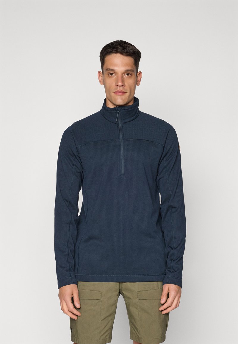 Marineblå zip-up fleece med lange ærmer, tekstureret stof, høj krave og kontrasterende syninger. Båret sammen med olivengrønne shorts.
