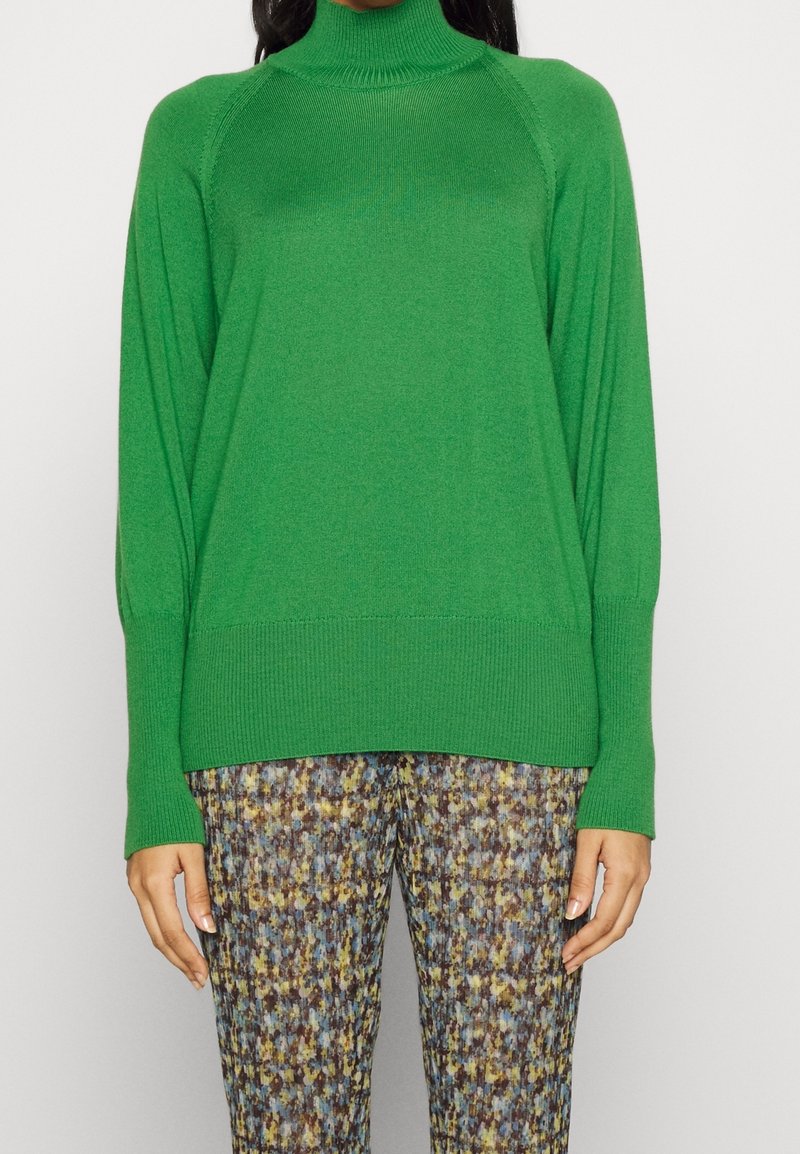 Maglione verde a collo alto lavorato a maglia con polsini e orlo a coste. Abbinato a pantaloni a fantasia in blu, marrone e senape.