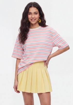 Jonge vrouw glimlacht, draagt gele stervormige oorbellen, een pastelgestreept kortemouwen top en een geplooide gele rok, staat met de hand op de heup.