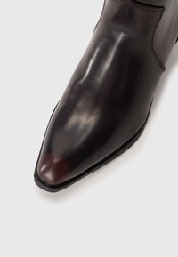Botte en cuir marron foncé au design épuré, à bout pointu, texture lisse et détails de couture minimaux. Semelle solide avec un léger éclat.