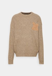 Béžový žebrovaný svetr s dlouhými rukávy, kulatým výstřihem a velkou oranžovou nášivkou ve tvaru písmena 'M'. Měkká textura a přiléhavý pas.