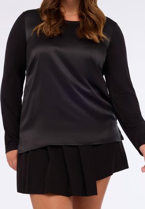 Frau, die ein langärmliges schwarzes Satintop trägt, kombiniert mit einem schwarzen, plissierten Minirock vor weißem Hintergrund.