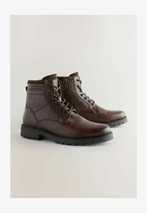 Bottes chelsea en cuir marron avec une texture lisse, bout rond, laçage à l'avant et fermeture éclair latérale pour un accès facile. Semelle en caoutchouc avec traction.