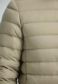 Pufferjack gemaakt van lichtbeige stof, met geborduurde secties met horizontale stiksels en een gladde, licht glanzende textuur.