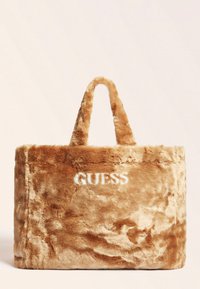 Beżowa torba tote z futra sztucznego o miękkiej fakturze, z podwójnymi uchwytami i białym logo "GUESS" z jednej strony.