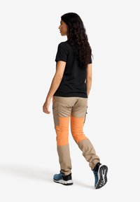 Svart t-shirt med kort ärm, beige baggybyxor med orange knäpatchar och blå löparskor. Håret är vågigt och faller förbi axlarna.