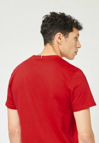 Rode katoenen t-shirt met korte mouwen. Heeft een ronde hals en een klein blauw, wit en rood label aan de achterkant. Glad textuur.