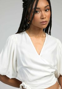 Femme avec des cheveux tressés portant des boucles d'oreilles en or et un chemisier blanc à manches bouffantes, regardant vers l'objectif sur un fond gris.