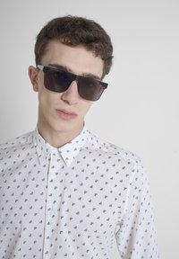 Camicia bianca con motivi geometrici neri, colletto a bottoni e maniche lunghe; modello che indossa occhiali da sole scuri con montatura quadrata.