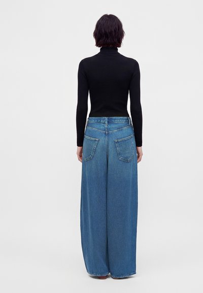 Personne portant un haut noir à manches longues et un jean bleu à jambes larges, debout de dos sur un fond clair uni.