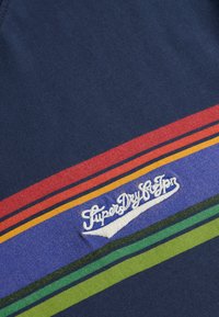 Tela azul oscuro con rayas horizontales en rojo, naranja, púrpura, verde y negro, con logo SuperDry bordado en blanco.