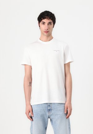 T-shirt bianco in cotone con colletto rotondo e maniche corte, caratterizzato da un piccolo logo sul lato sinistro del petto e una vestibilità rilassata. Indossato con jeans azzurri chiari.
