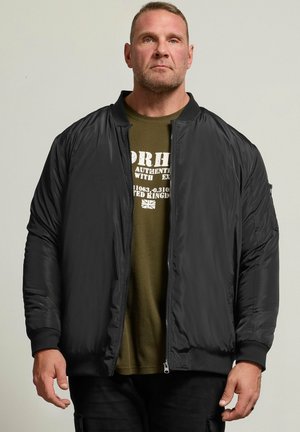 Hombre con una chaqueta bomber negra sobre una camiseta verde oliva con texto blanco y una pequeña bandera del Reino Unido.
