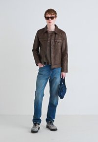 Smeđa kožna jakna preko sive majice, plave traperice i sive cipele. Drži tamnoplavu torbu, nosi naočale za sunce, stoji naslonjen na svijetlu pozadinu.