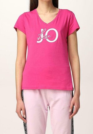 Kobieta ubrana w jasnoróżowy T-shirt z dekoltem w serek z tekstem "JLO" na białych i jasno-różowych spodniach z czarnymi wzorzystymi paskami po bokach.