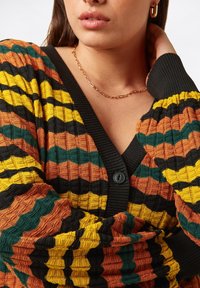 Bunt gestreifter Cardigan mit einem strukturierten Muster in Orange, Gelb, Grün und Schwarz; Knopfdetails auf der Vorderseite; gerippte Bündchen.