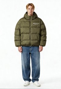 Giacca imbottita verde oliva con design trapuntato, cerniera frontale e cappuccio. Paio di jeans blu a gamba larga e sneakers bianche.