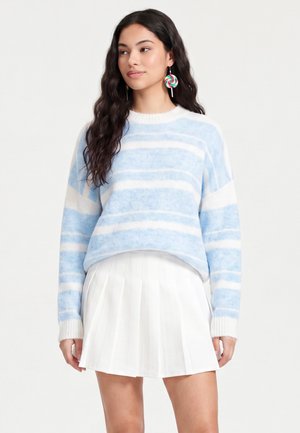 Jeune femme aux longs cheveux foncés porte un pull rayé bleu et blanc, une jupe plissée blanche et des boucles d'oreilles colorées en forme de sucette.