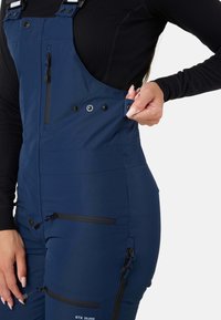 Marinblå vattentäta overaller med justerbara remmar, flera fickor med dragkedja och en slät yta med kontrasterande svarta detaljer.