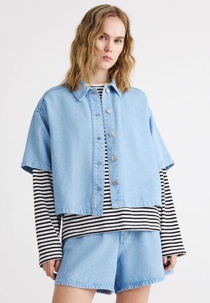 Junge Frau mit welligem blondem Haar trägt eine hellblaue Jeansjacke mit kurzen Ärmeln, ein schwarz-weiß gestreiftes Langarmshirt und Jeansshorts.