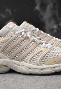 Weiße und beigen Adidas Laufschuhe mit Mesh-Design und drei charakteristischen Streifen, in einer Nahaufnahme seitlich auf einer grauen Oberfläche dargestellt.