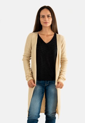 Femme avec de longs cheveux bruns portant un cardigan beige à côtes, un t-shirt noir en V et un jean bleu, se tenant devant un fond blanc.