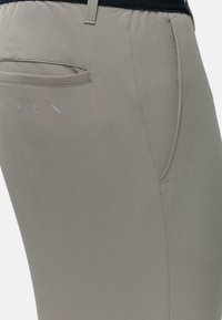 Un pantalon gris clair présente un design ajusté avec une texture lisse, une poche arrière et un logo en matériau réfléchissant.