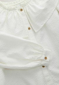 Weißes, strukturiertes Blusenshirt mit Rüschenausschnitt, Holzknöpfen und gerafften Ärmeln. Der Stoff hat ein dezentes, geprägtes Muster.