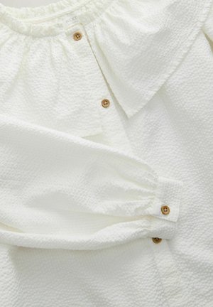 White textured blouse with a ruffled collar, featuring wooden buttons and gathered sleeves. The fabric has a subtle embossed pattern.