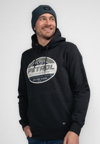 Sudadera negra con un gran gráfico circular que presenta montañas y el texto "PATROL". Combinada con un gorro azul oscuro, exhibida sobre un fondo neutro.