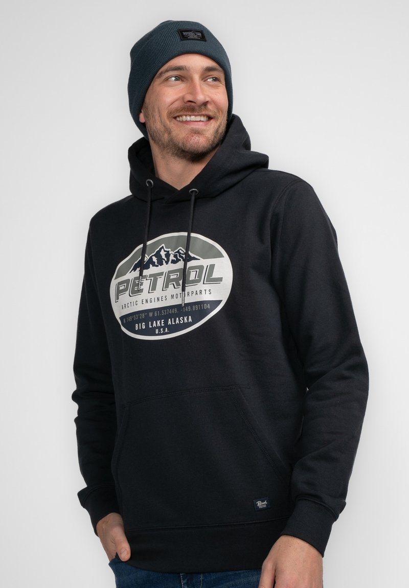 Sudadera negra con un gran gráfico circular que presenta montañas y el texto "PATROL". Combinada con un gorro azul oscuro, exhibida sobre un fondo neutro.