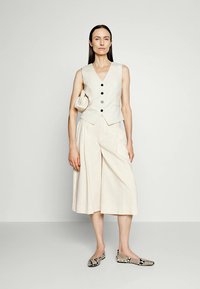 Beige sleeveless vest dengan kancing hitam, dipadukan dengan kulot lebar berwarna terang. Aksesori termasuk tas kecil beige dan sepatu datar bermotif.