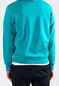 Petrolfärgad bomullströja med huva, med ribbad midja och ärmslut, sedd bakifrån, i kombination med en vit undertröja.