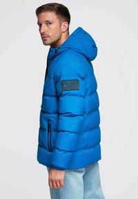 Sininen toppatakki hupulla, jossa tikattu pehmuste, mustat vetoketjut ja vasemmassa hihassa oleva merkki. Sileä, kiiltävä kangaspinta.