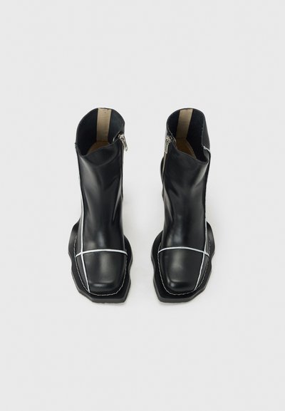 Μαύρα δερμάτινα μποτάκια αστραγάλου με λευκή αντίθετη ραφή, τετράγωνα άκρα, πλαϊνά φερμουάρ και χοντρές σόλες σε απλό ανοιχτό φόντο.