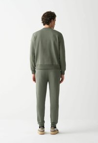 Hanorac verde și pantaloni jogging asortați, fabricați dintr-un material moale, având mânșete ribelate și o croială relaxată. Adidașii bej completează ținuta.